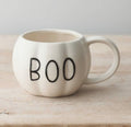 An off-white ceramic mug shaped like a small pumpkin with the word 'BOO' printed in black on the front, sitting on a light wooden surface against a neutral gray background.