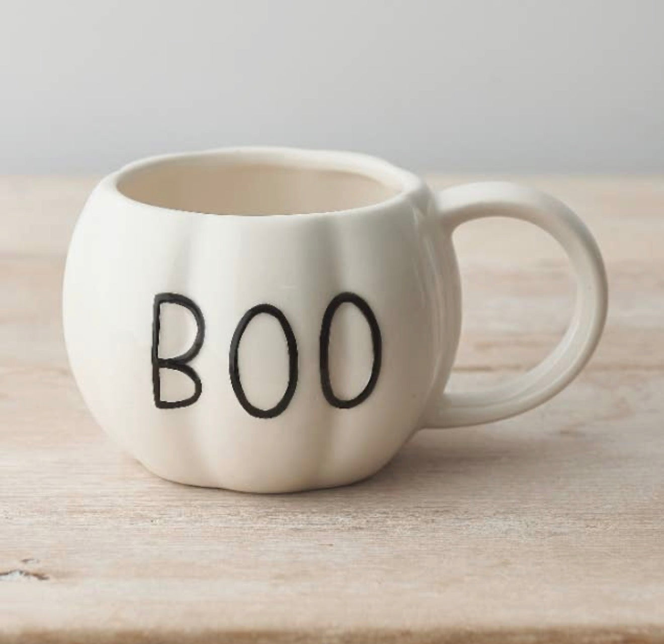 An off-white ceramic mug shaped like a small pumpkin with the word 'BOO' printed in black on the front, sitting on a light wooden surface against a neutral gray background.