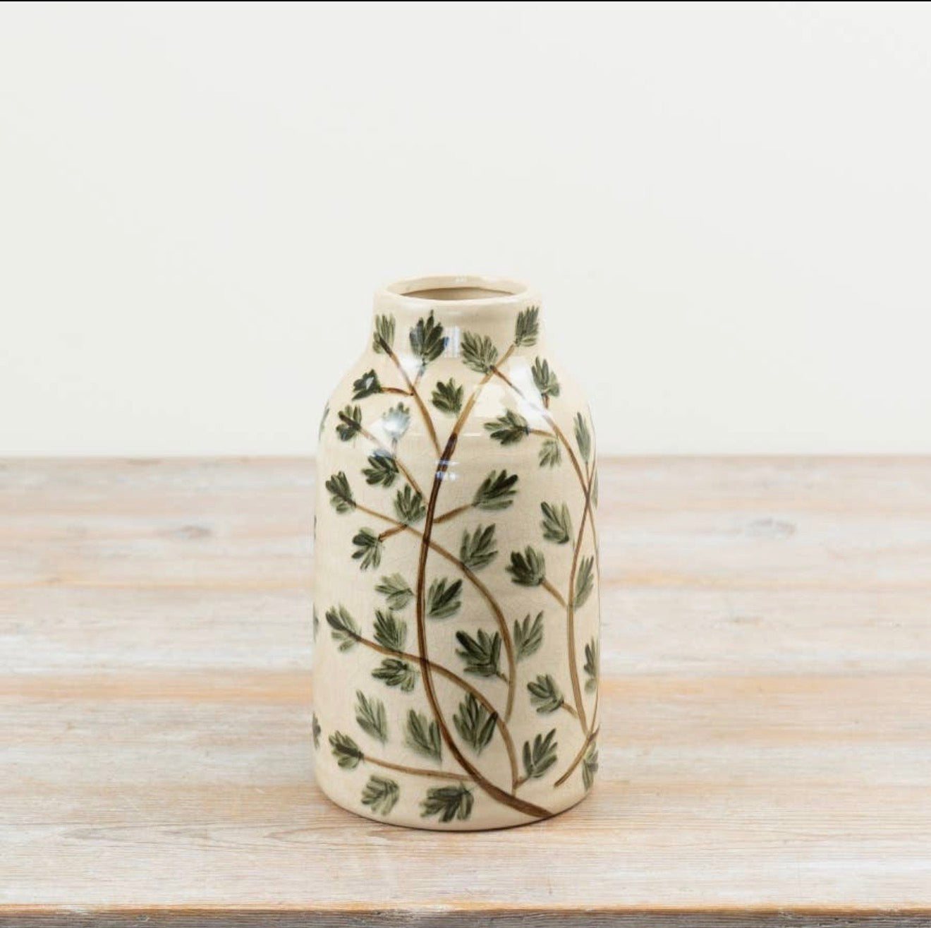 A small ceramic vase with a cream-colored glaze, decorated with a hand-painted green leafy vine pattern, set on a light wooden surface against a white background.