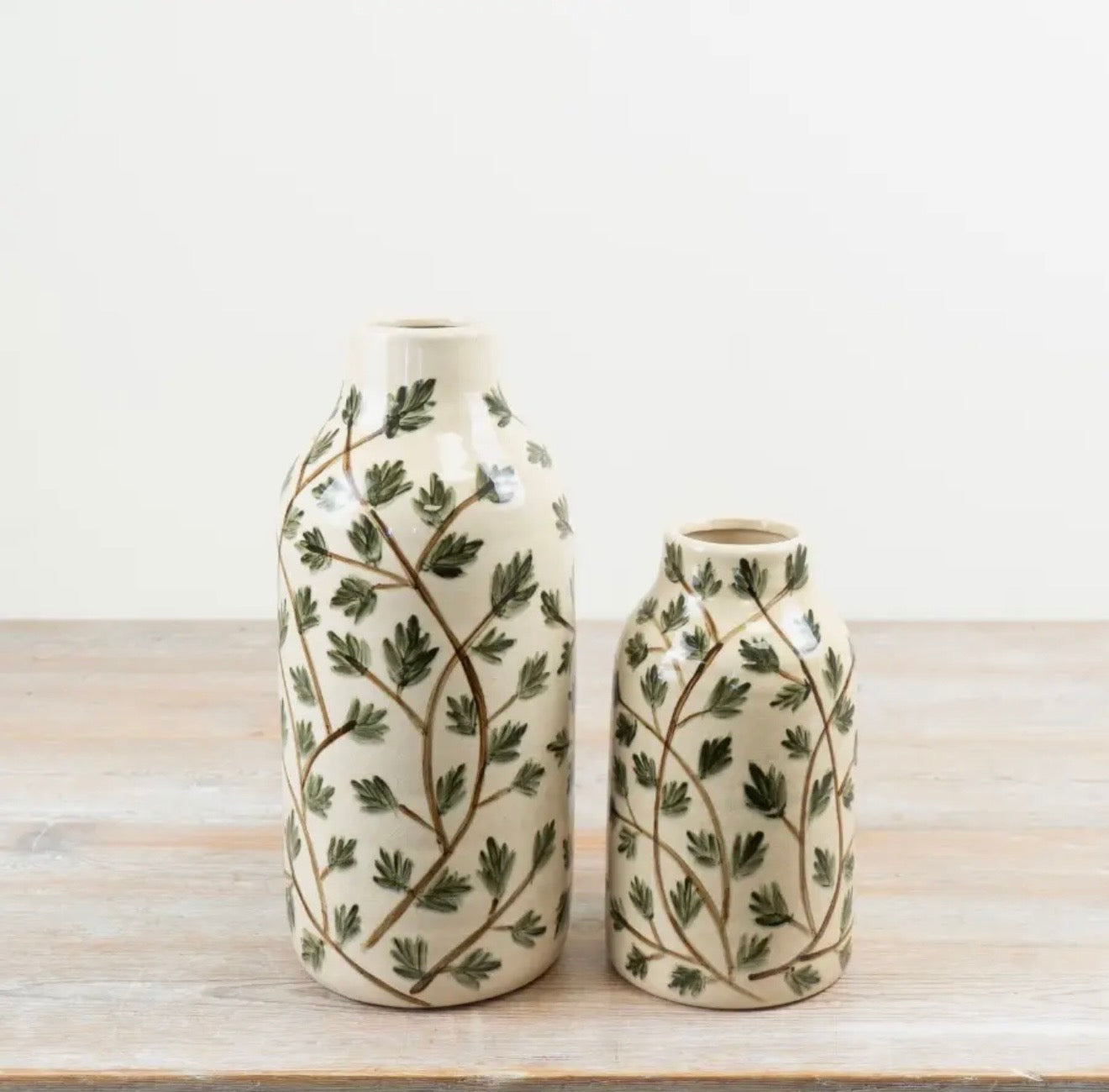 Two ceramic bottles of different sizes with a green leafy vine pattern on a cream background, displayed on a light wooden surface against a white wall.