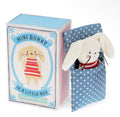 Mini white fabric bunny wearing a red striped outfit tucked into a blue polka dot bed beside its illustrated gift box.